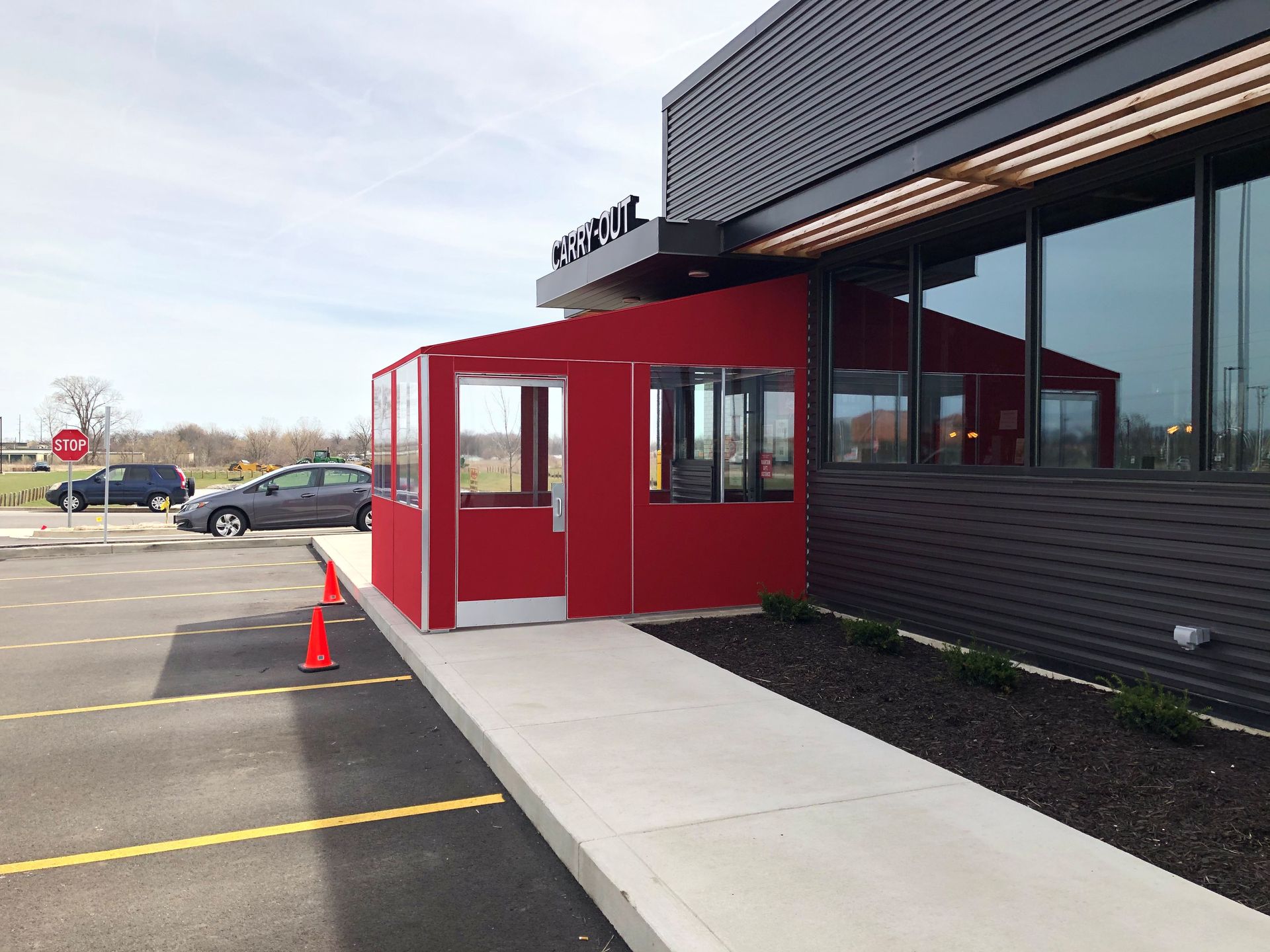 Red entrance canopy and glass door at a modern building, beside a parking lot and sidewalk.
