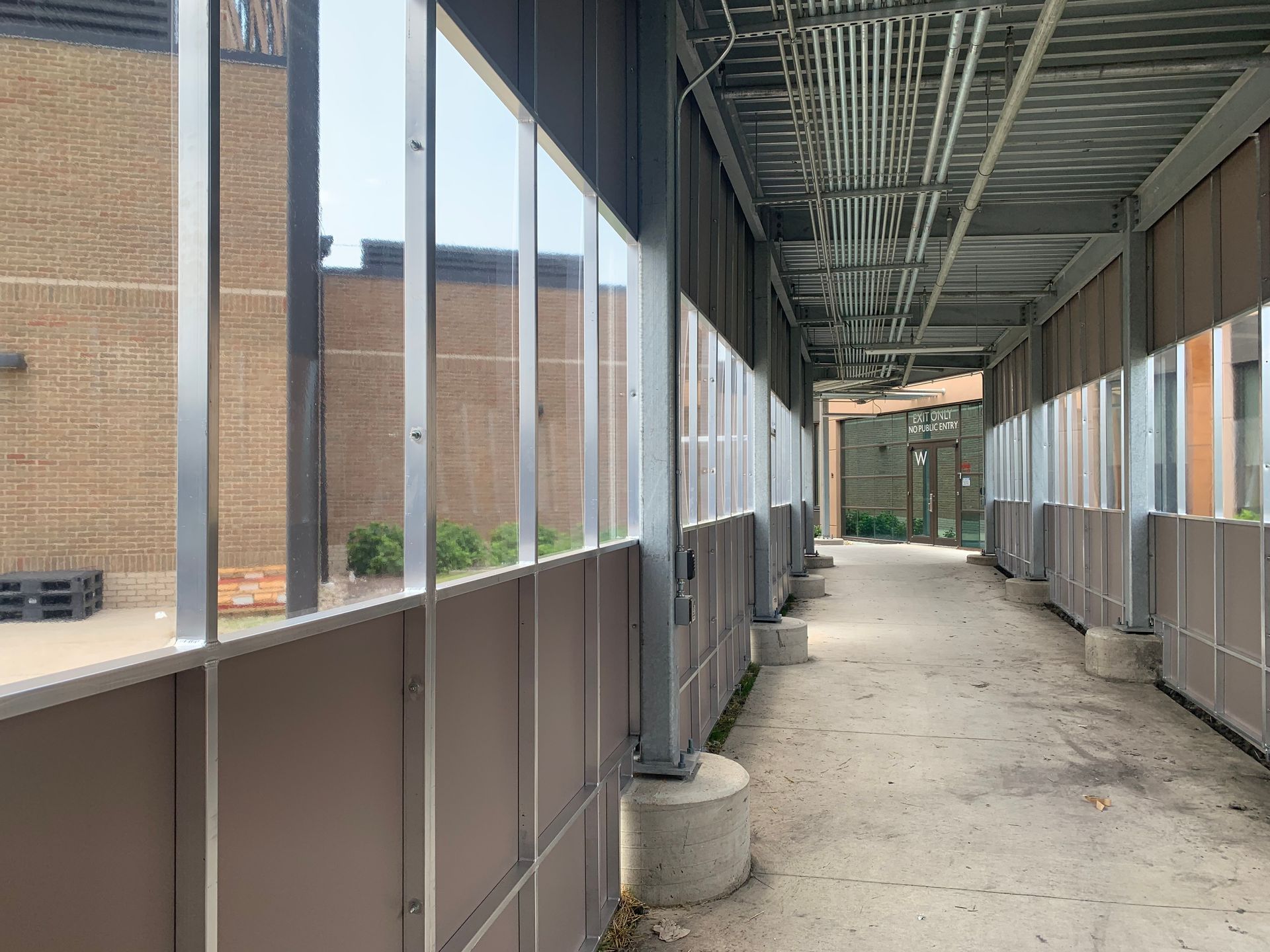 Covered walkway with metal framing and translucent panels beside a brick building, under construction