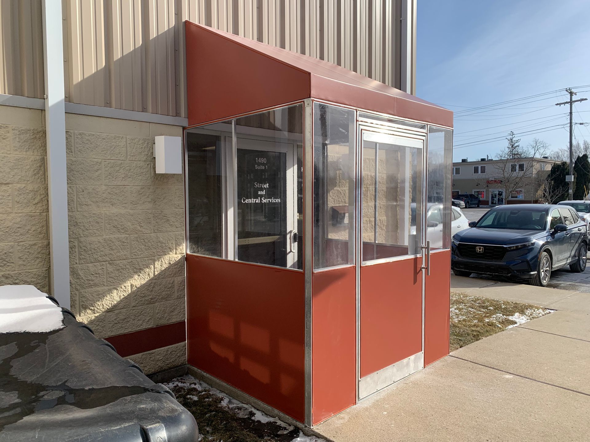 Red storefront kiosk with glass windows beside a sidewalk and parked car