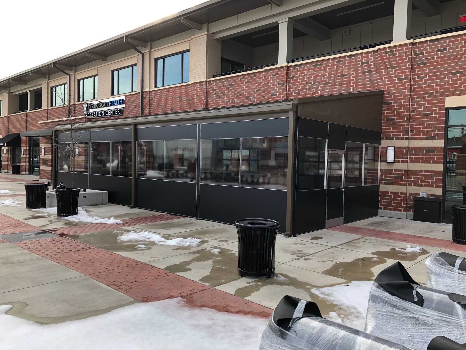 Outdoor brick patio with a black glass-walled enclosure and trash cans in front of a building