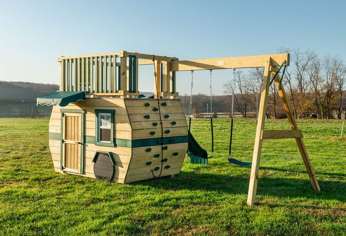 A wooden playhouse with a swing set and climbing wall is sitting in the middle of a grassy field
