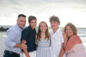 Family of five smiling on a beach; sunny, overcast sky.