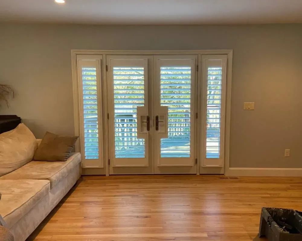 Living room with wooden floors, light gray walls, a tan couch, and white French doors with shutters.