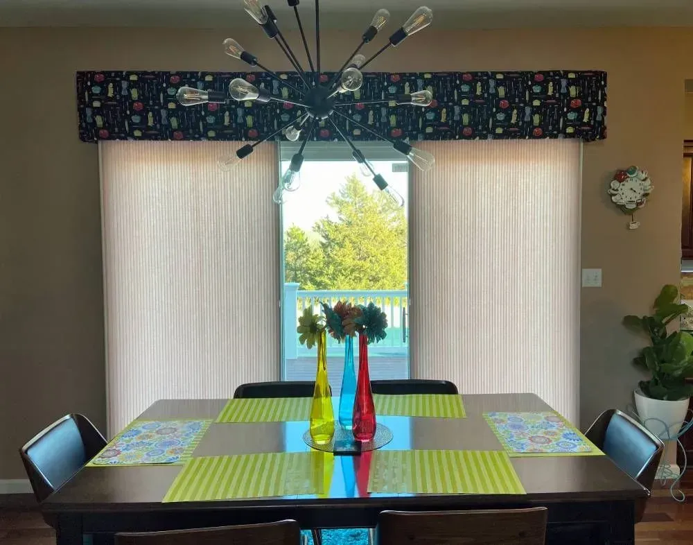 Dining room with table set below window with light-colored curtains and patterned valance. A chandelier hangs above the table.