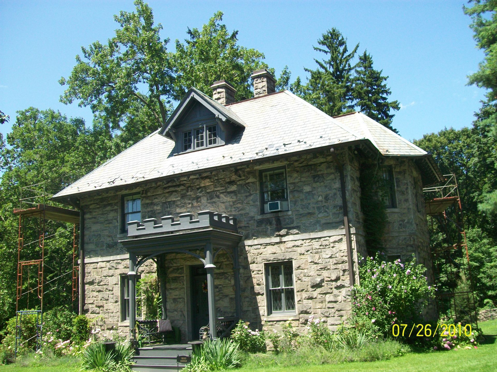 Stone house with a white roof and porch, surrounded by tall green trees under a clear blue sky.