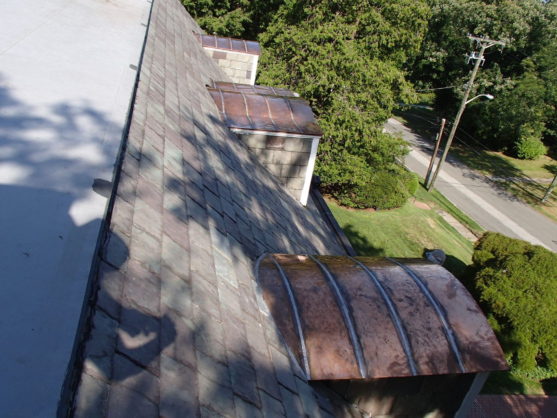 Rooftop with shingles and two small metal awnings beside a tree-lined street