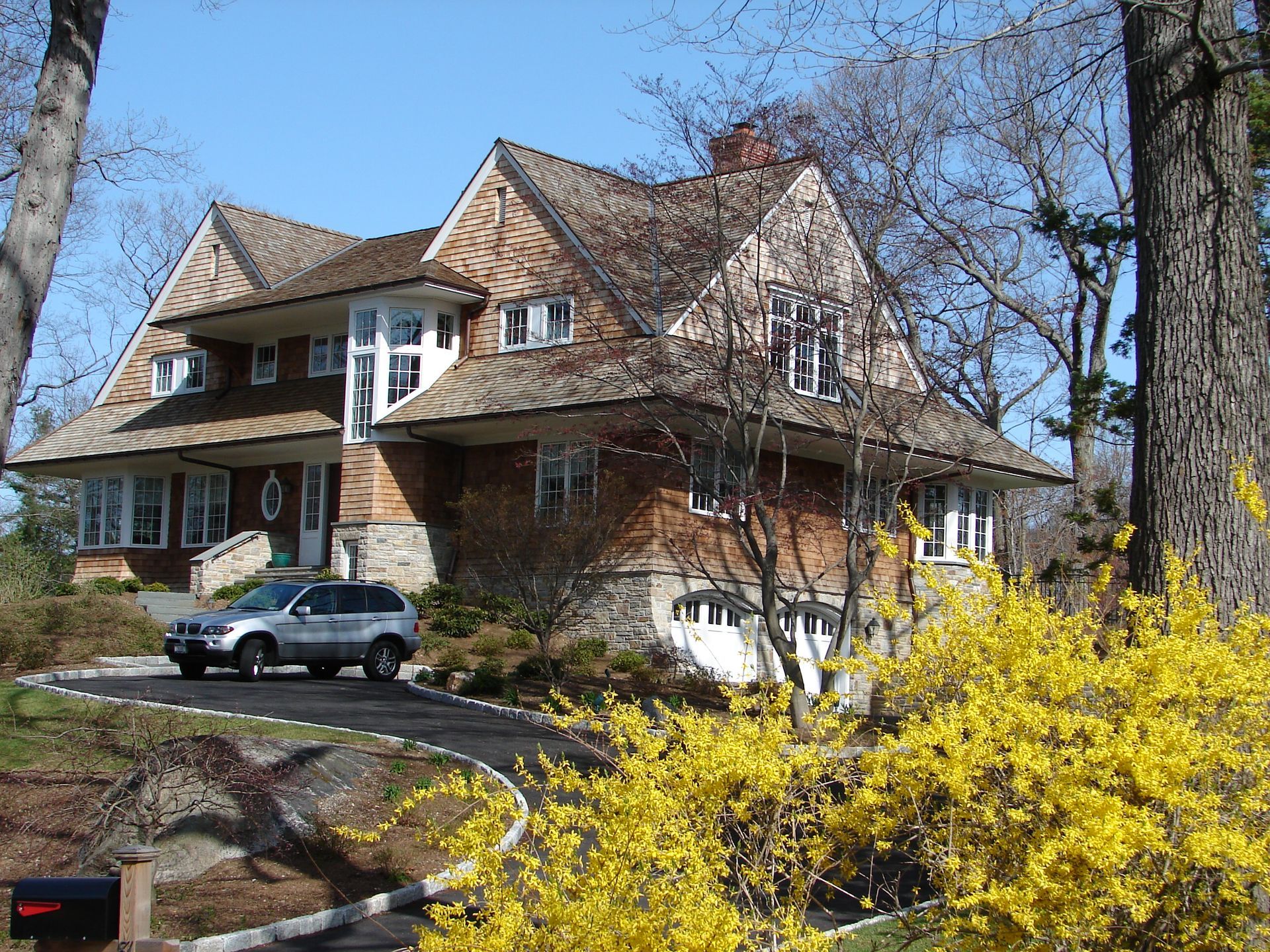 Large house with a steep roof, parked car, and bright yellow shrubs in a sunny yard