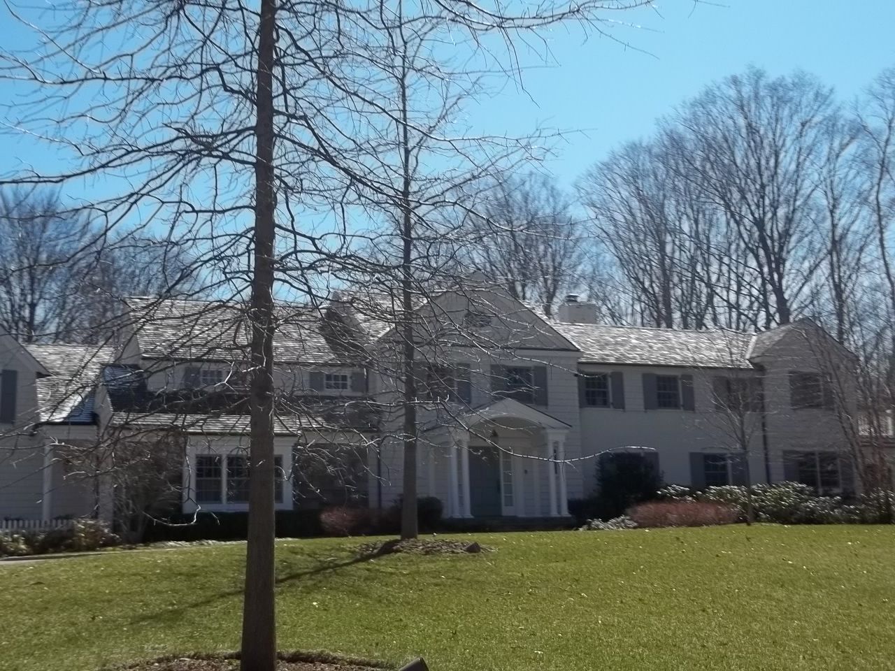 Large stone house with white trim behind a grassy lawn and leafless trees on a sunny day
