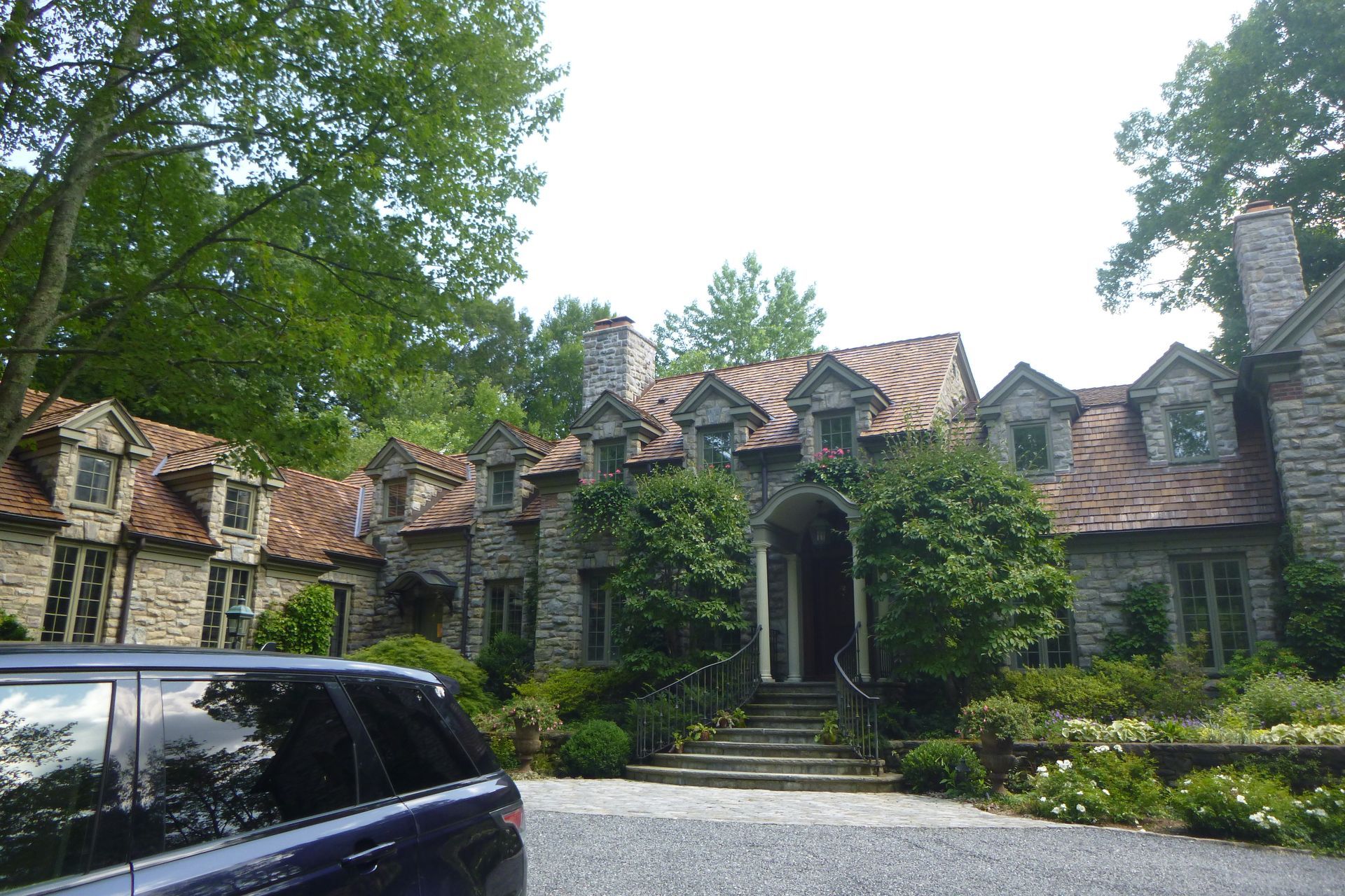 Stone mansion with red-tiled roofs behind trees, with a black car parked in front