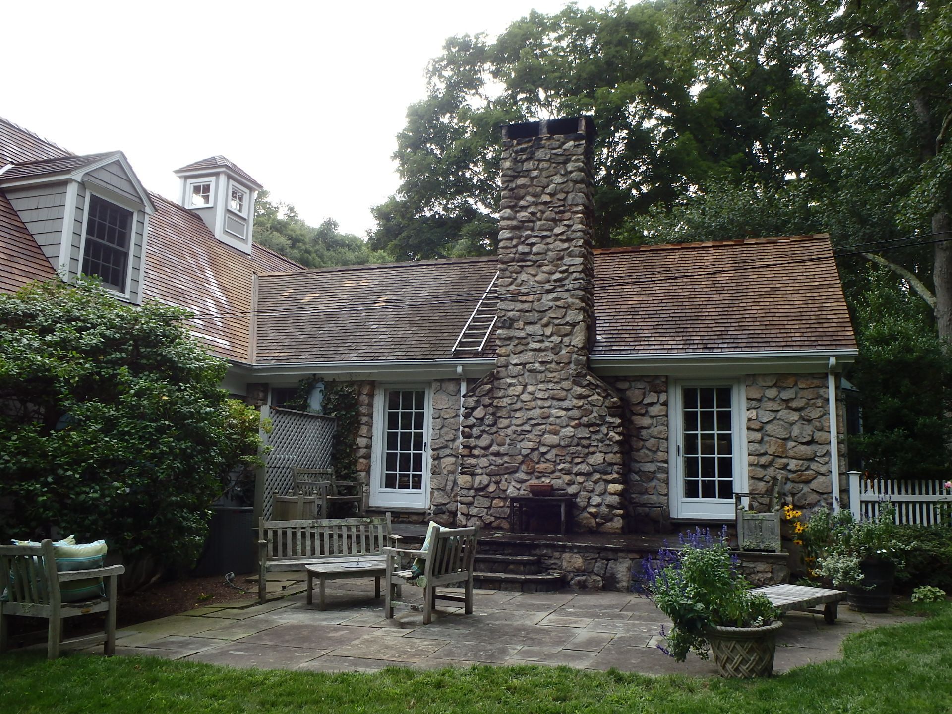 Cottage with stone chimney, shingled roof, and small patio surrounded by lush greenery