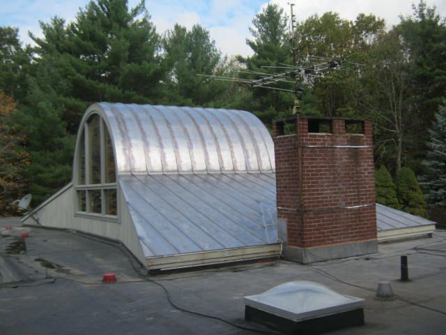 Curved metal-roofed building with brick chimney on a rooftop, surrounded by trees and sky.