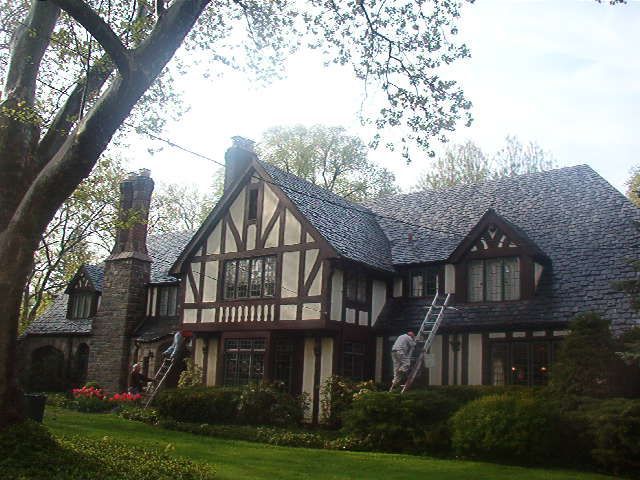 Large Tudor-style house with dark roof, white trim, and a ladder leaning against the front in a leafy yard
