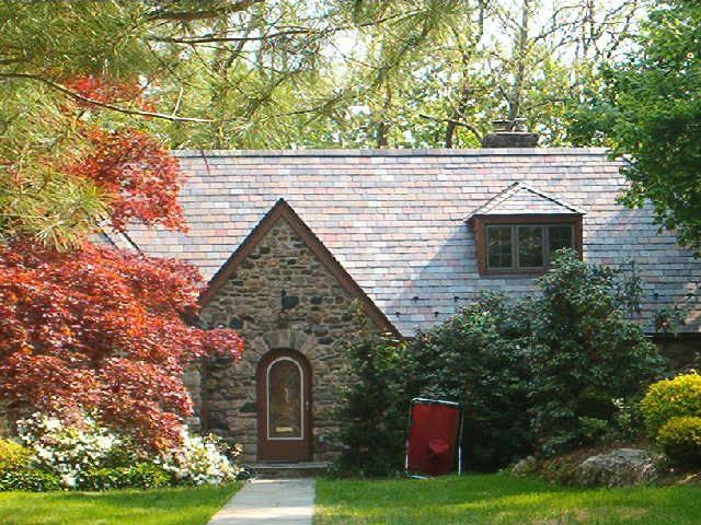 Stone cottage with a shingled roof, framed by red and green trees in a garden.