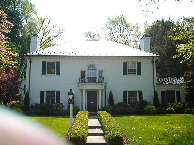 White two-story house with a centered front door, balcony, and manicured lawn in a sunny yard