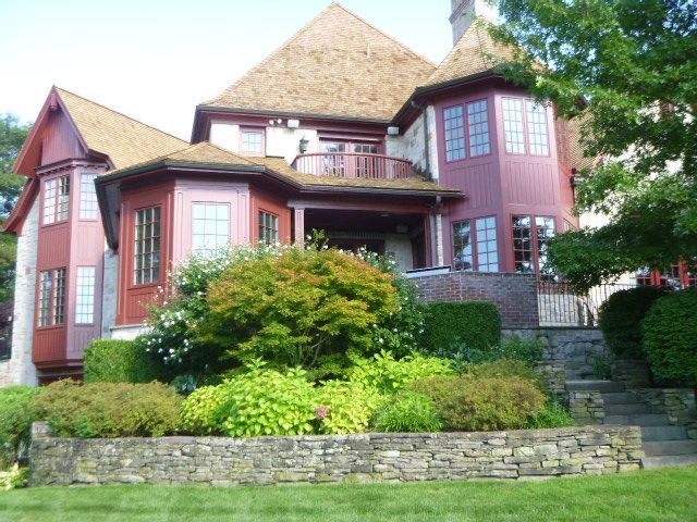 Large red Victorian-style house with bay windows, stone retaining wall, and lush green landscaping