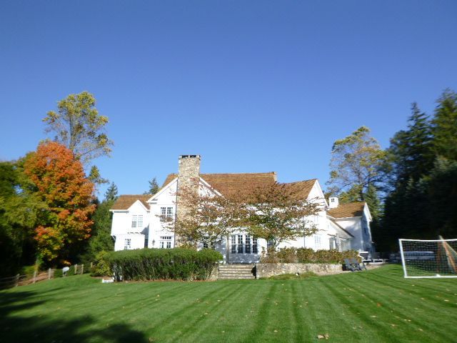 White house with a stone chimney on a green lawn under a clear blue sky, framed by autumn trees