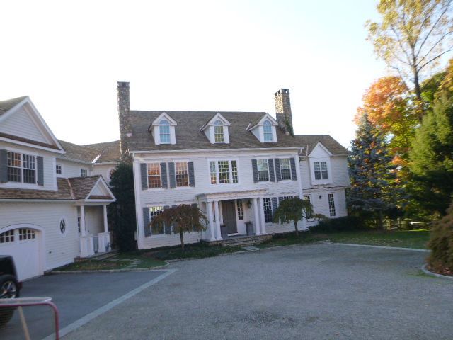 Large white colonial-style house with a front porch, chimneys, and a driveway, surrounded by trees