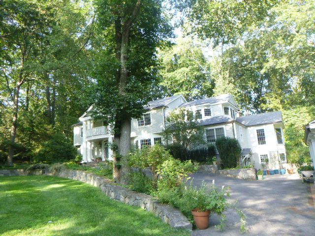 White house with porch and driveway surrounded by tall trees and greenery