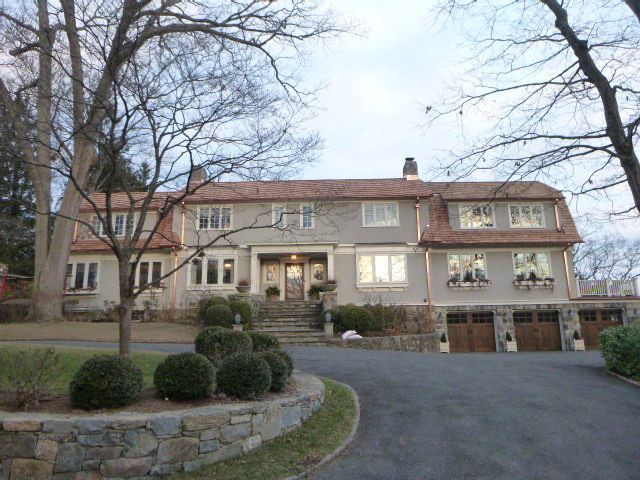 Large stone house with a red roof, lit windows, and a driveway framed by bare trees and landscaping