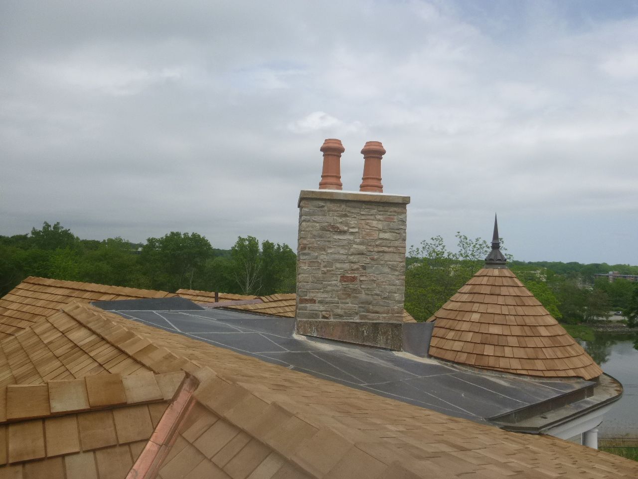 Rooftop with brick chimney, tan shingles, and a cloudy sky over trees in the distance