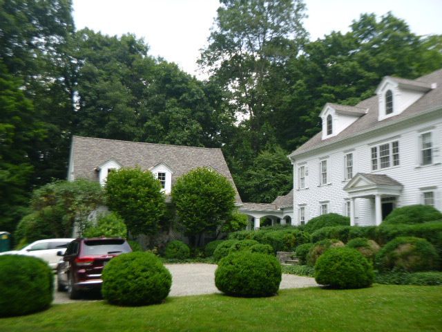 Suburban home with a white house, detached garage, parked SUV, and manicured shrubs under trees