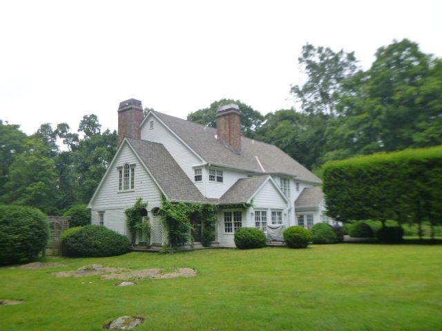 White cottage-style house with two chimneys, surrounded by lush green lawn and trees.