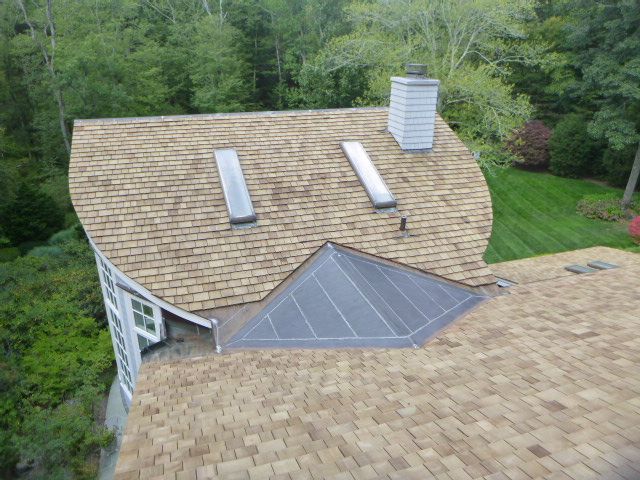Aerial view of a shingled roof with skylights and a chimney, surrounded by trees.