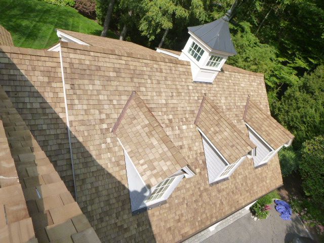 Aerial view of a tan shingled house roof with dormer windows and a small cupola, surrounded by trees.