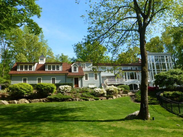 Large white house with red roof on a green lawn, framed by trees under a clear blue sky