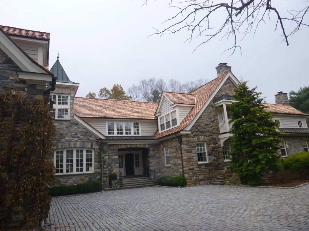 Stone house with red-tiled roof and ivy, seen from a cobblestone driveway under an overcast sky