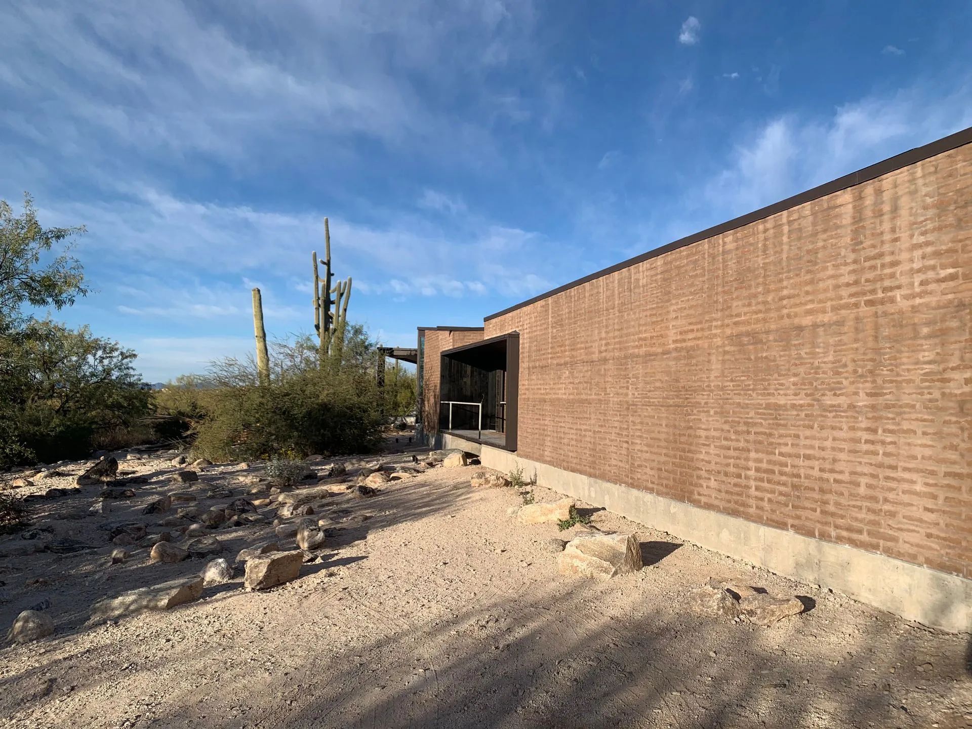 Desert building with tan brick wall, rocky ground, and tall cacti under a blue sky