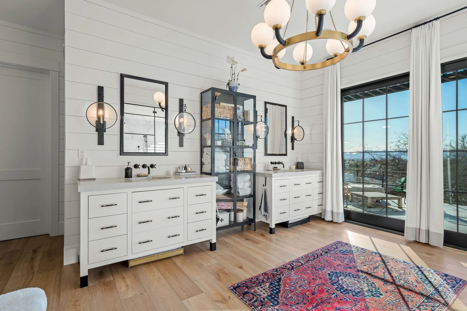 Bright bedroom with white dressers, mirrored wardrobes, chandelier, and patterned rug by large windows.