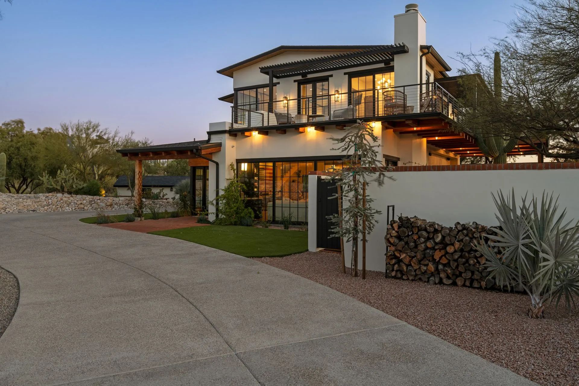 Modern two-story house with warm lights, stone driveway, and desert landscaping at dusk