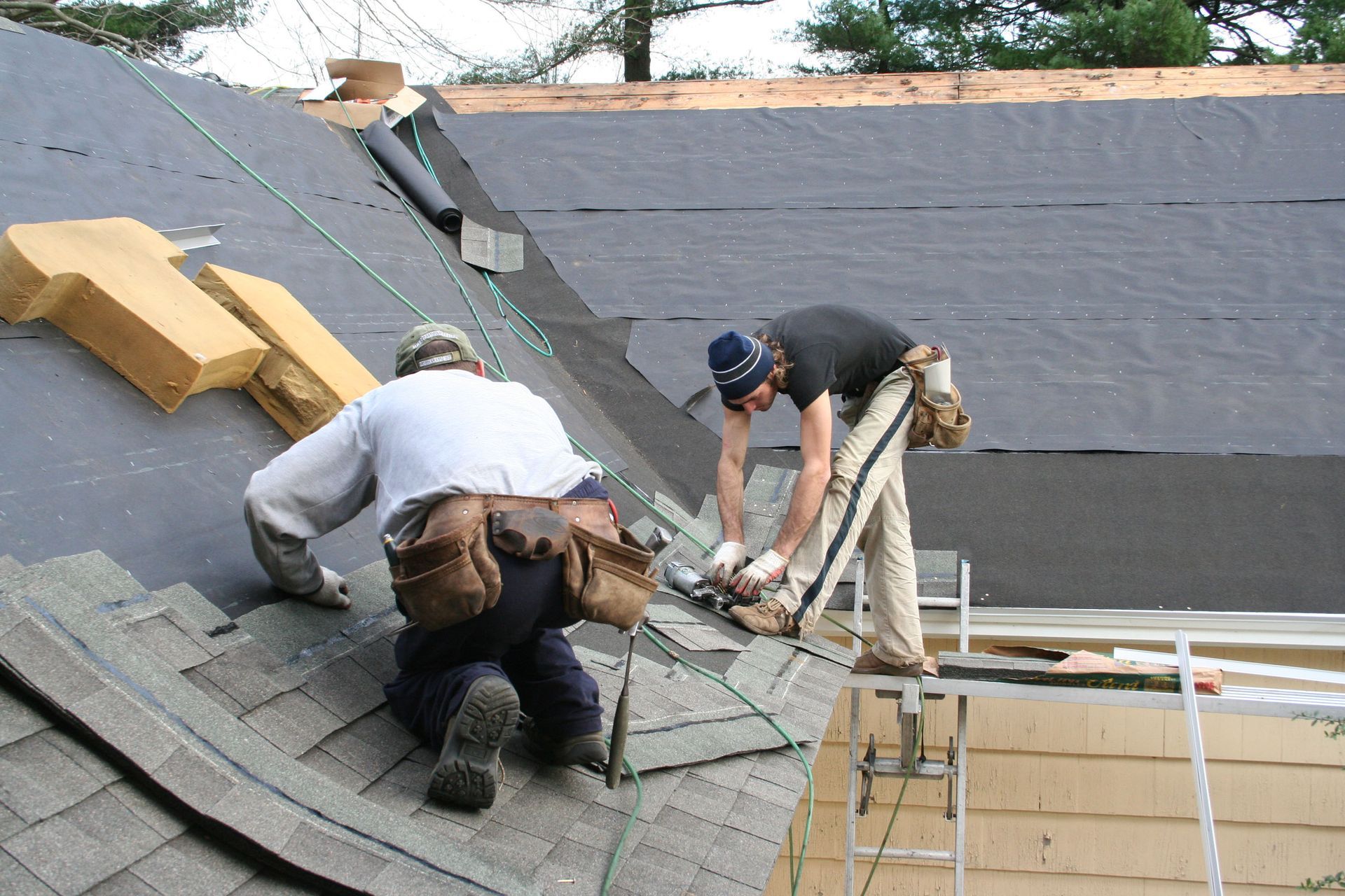 Two roofers working on a roof, installing shingles. One kneels, the other stands, using a tool.