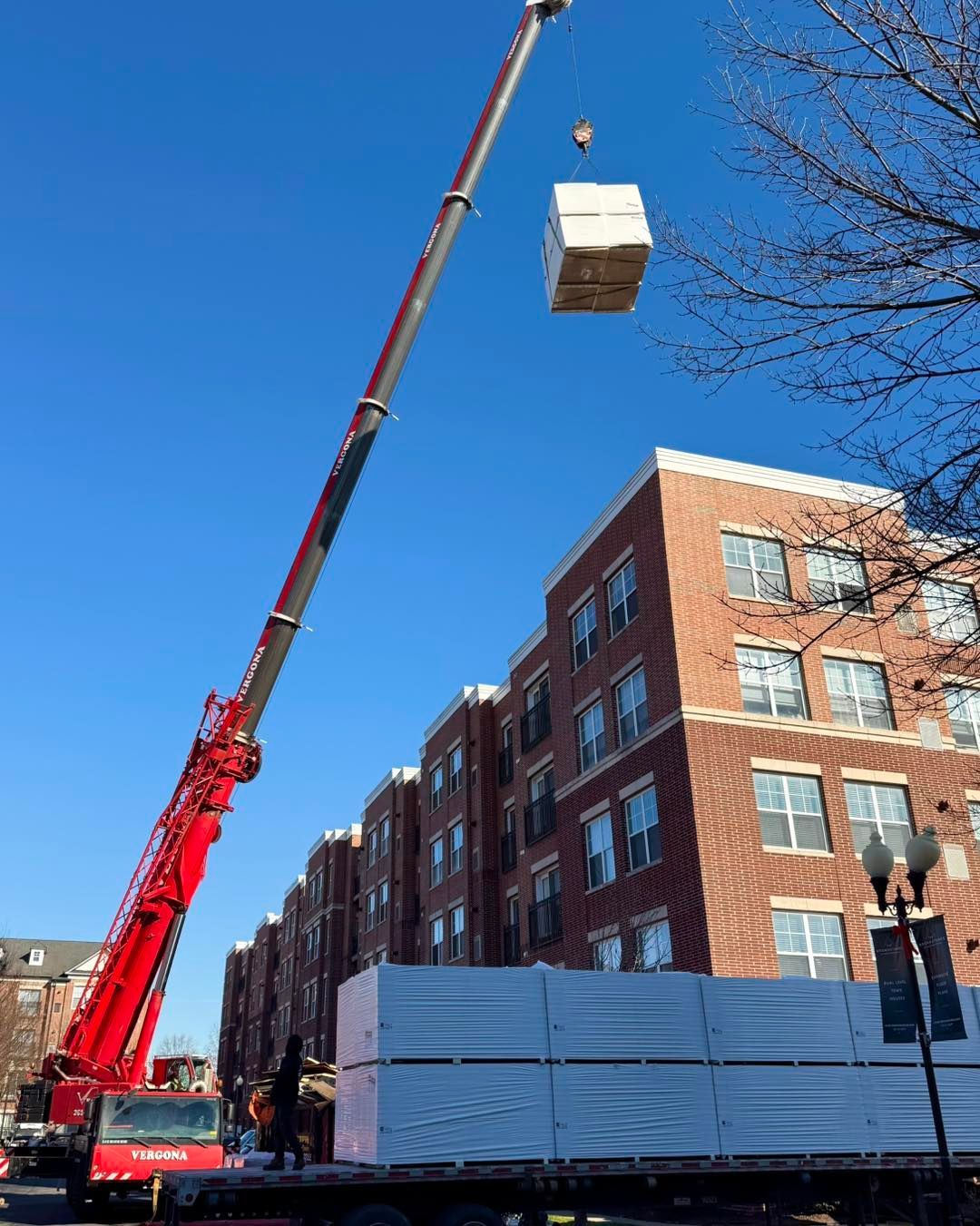 A large crane lifts a box towards a brick building. Workers near a truck hold white panels. Blue sky.