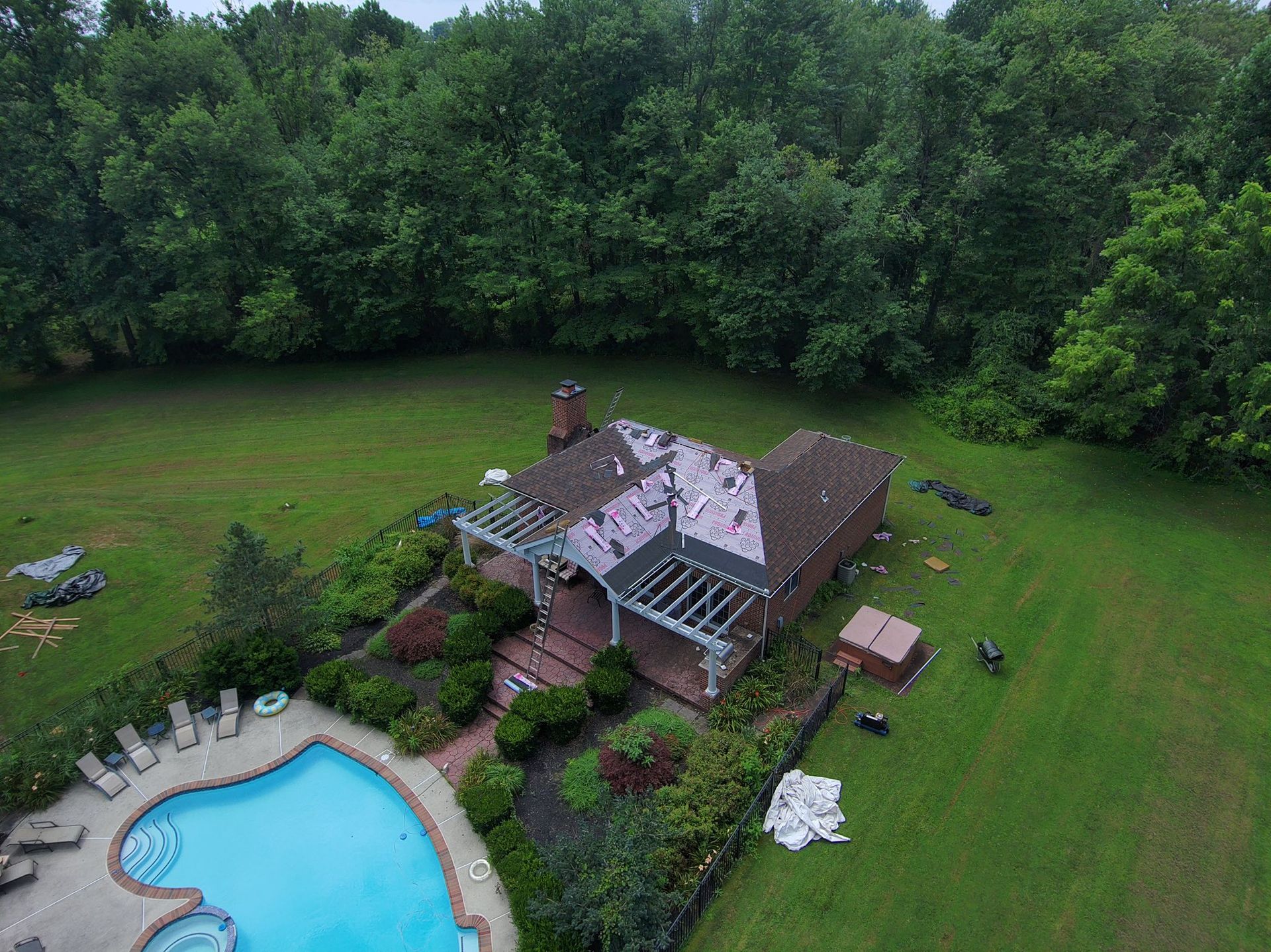 Aerial view of a house with pool, yard, and surrounding trees. Roofing work is in progress.
