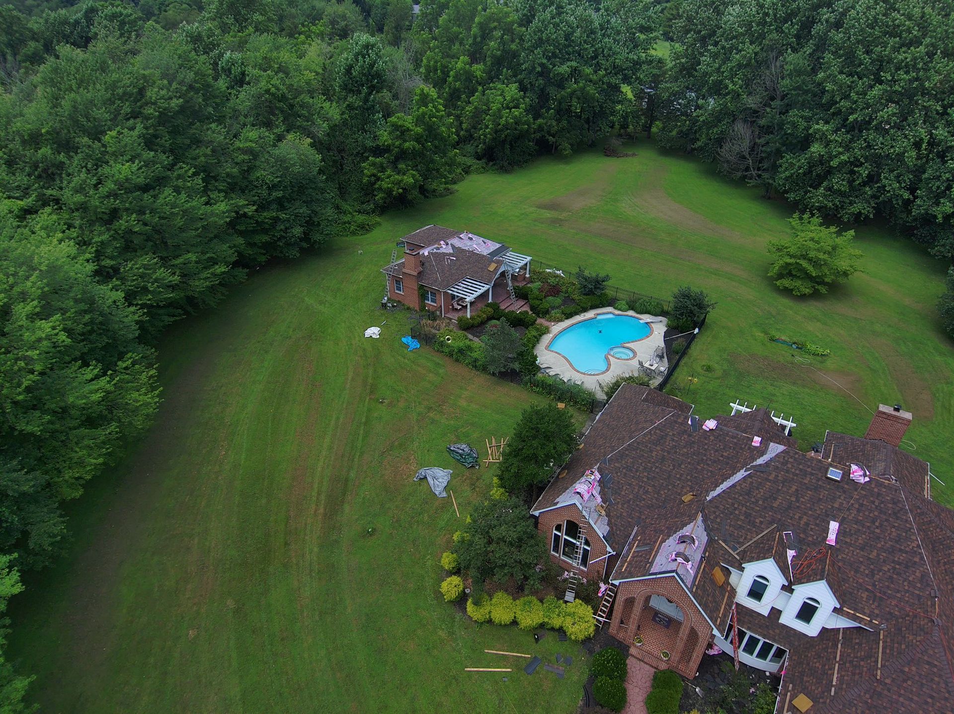 Aerial view of a house with a pool and smaller adjacent building on a grassy hill surrounded by trees.