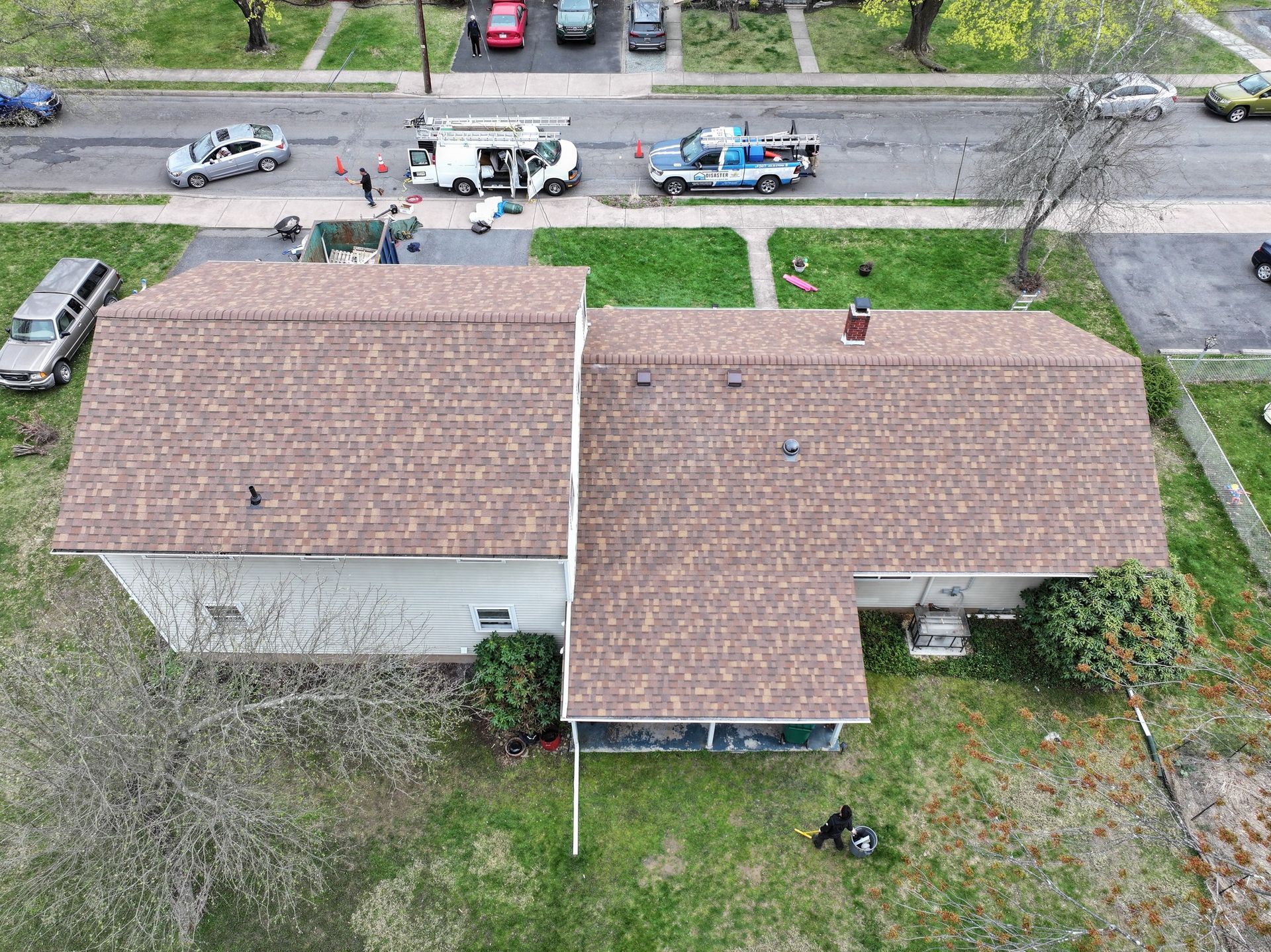 Aerial view of a house with brown roof and cars parked on the street. People and vehicles appear to be working in the street.