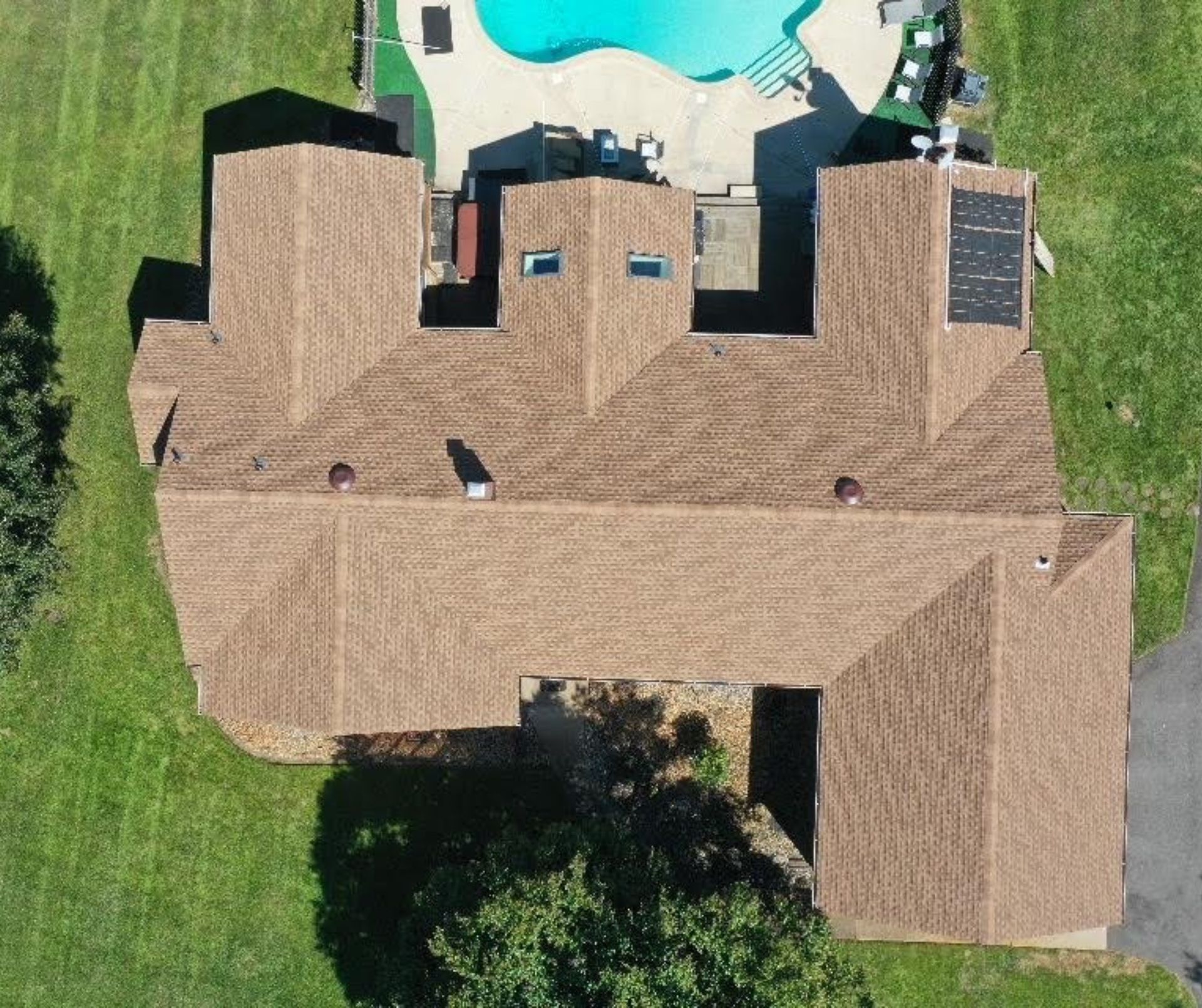 Aerial view of a brown-shingled house with a pool and solar panels on the roof, surrounded by green grass.