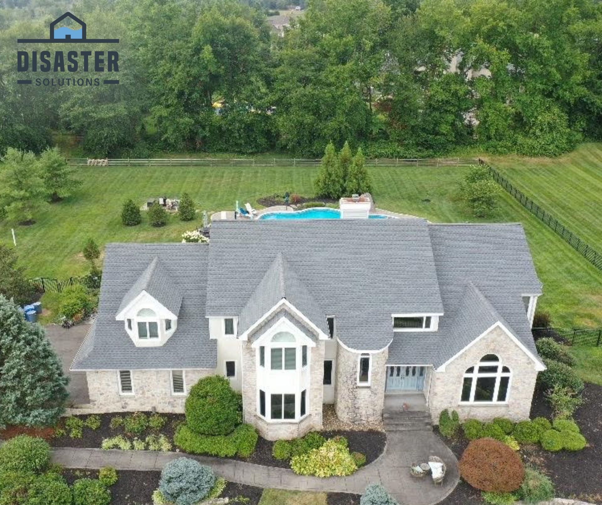 Aerial view of a large house with a gray roof and a swimming pool in the backyard, surrounded by green trees and grass.