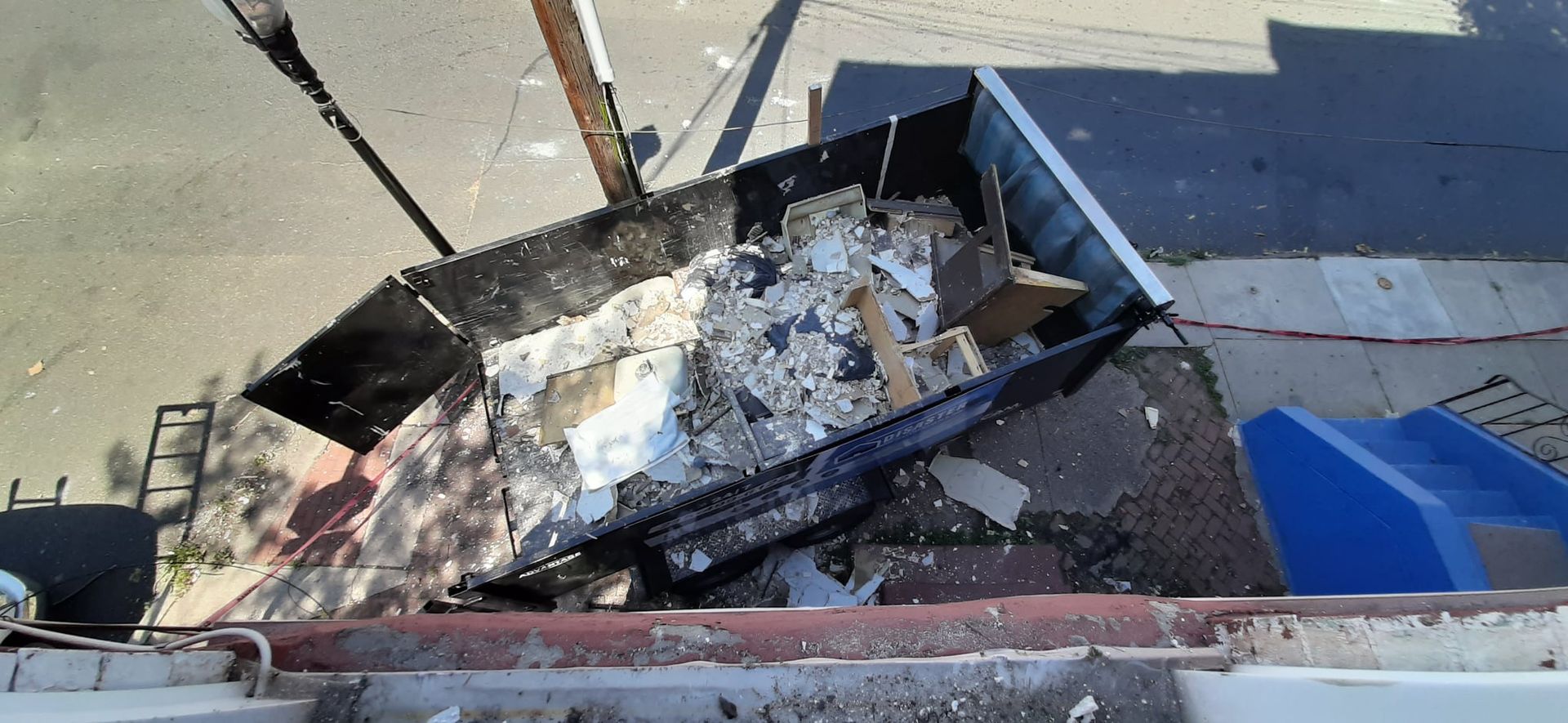 A black dumpster overflowing with debris sits outdoors on a sunny day.