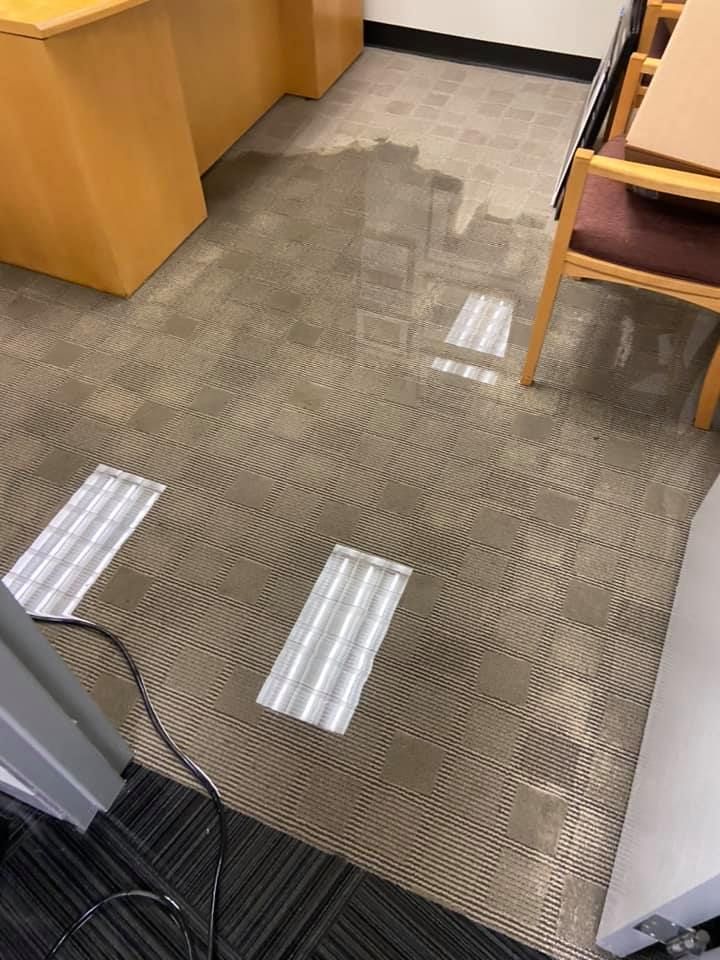 Water damage floods office carpet. Desk and chairs are visible. Two light fixtures lay on floor.