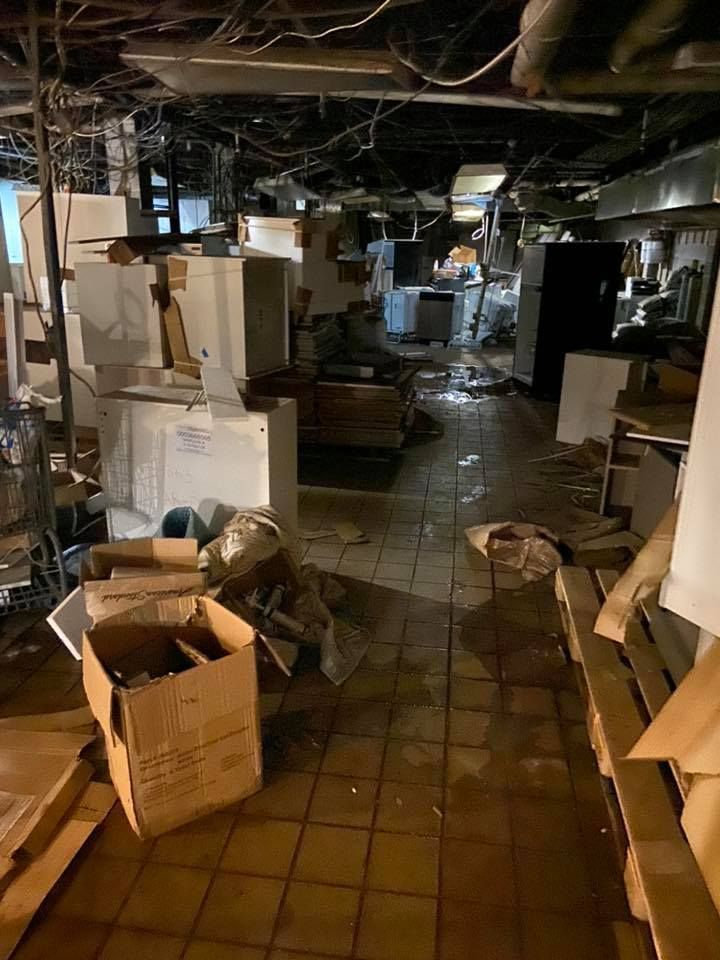 Cluttered basement room with appliances, boxes, and wet floor; possibly damaged from flooding.