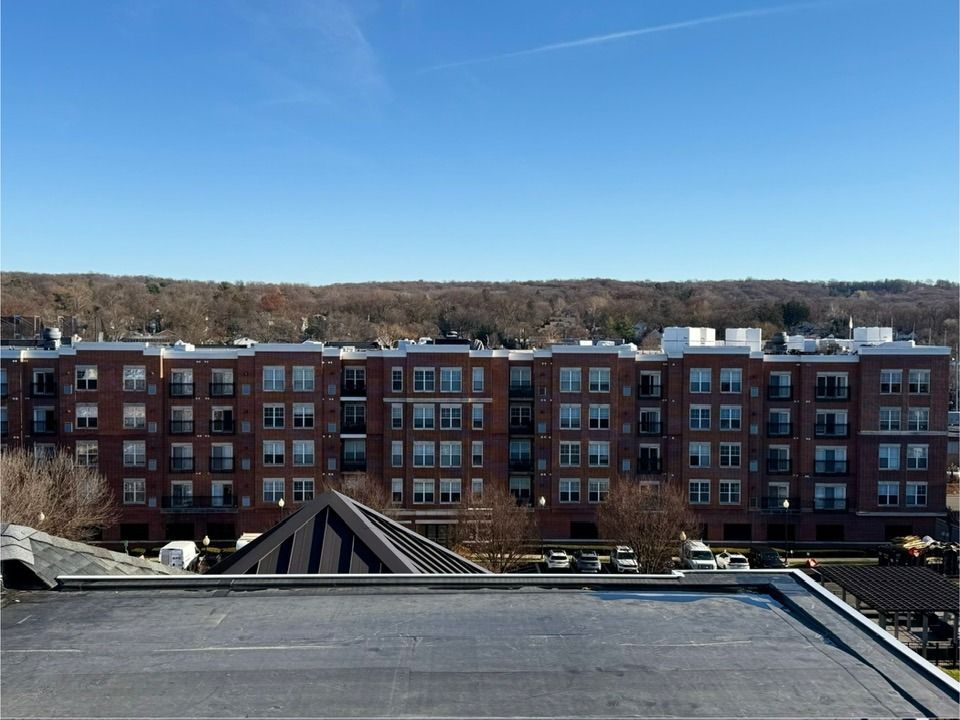 Multi-story brick apartment building on a sunny day. Forest visible in the background, blue sky.