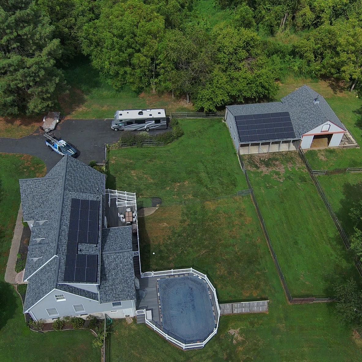 Aerial view of a house, barn, and RV surrounded by green grass and trees; solar panels on roofs.