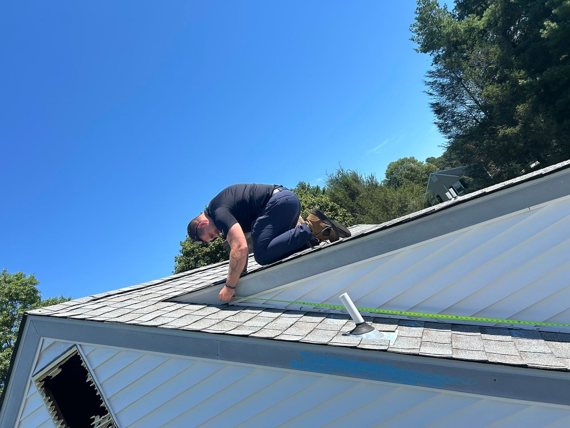 Man on a roof, inspecting shingles on a sunny day. He is crouching near the edge, wearing dark clothes.