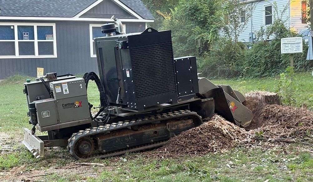 Black skid-steer excavator pushing dirt in a yard beside a blue house and trees