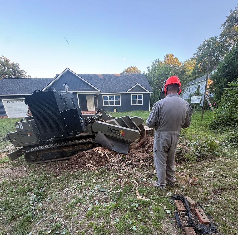 Excavator demolishing debris in front of a gray house; worker in orange hard hat stands nearby.