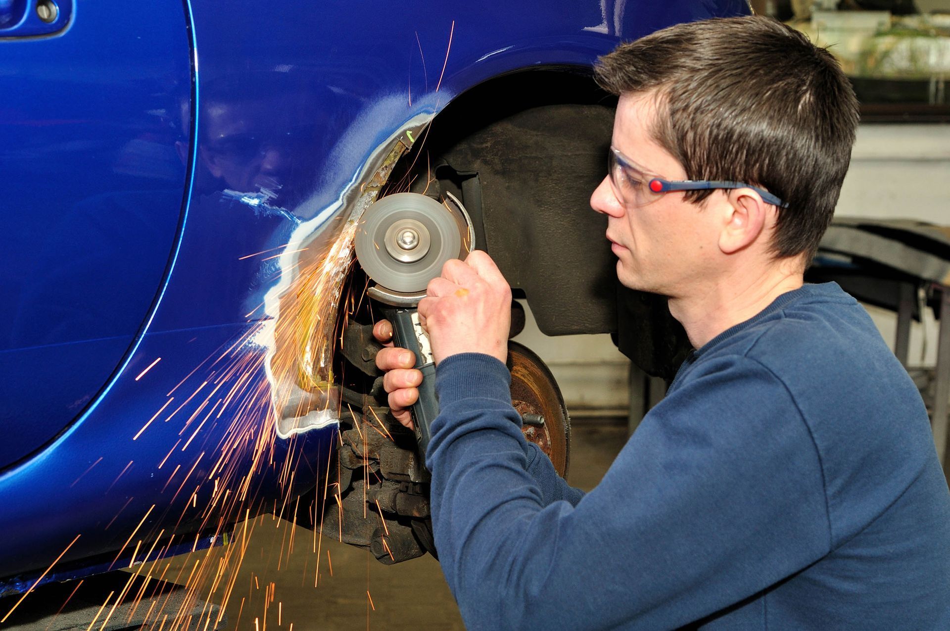 Person using a power grinder on a blue car wheel well, with sparks flying.