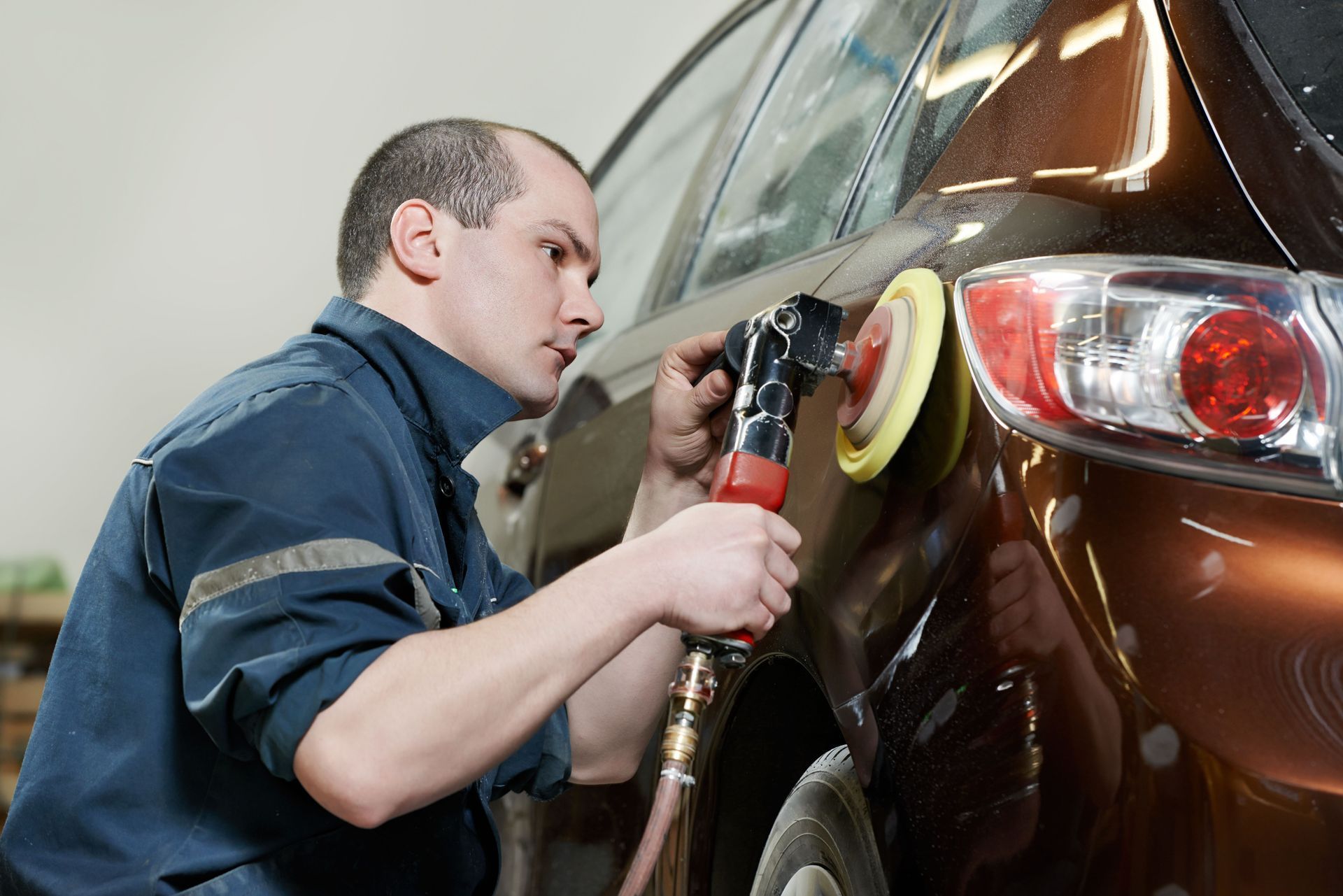 Mechanic polishing a brown car’s rear fender with a power buffer in a garage